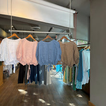 Load image into Gallery viewer, Aurora Boutique Miami Delmar linen blouses in white, pink, blue, and tan hanging on a rack in a clothing store.