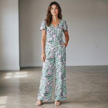 Load image into Gallery viewer, Model wearing an Aurora Boutique Miami Cite floral jumpsuit with green palm leaves on a light pink background