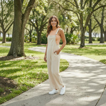 Load image into Gallery viewer, Model wearing Aurora Boutique Miami beige linen wide leg jumpsuit with a peach tank top in a park setting
