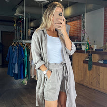Load image into Gallery viewer, Aurora Boutique Miami beige linen shirt dress with button front, breast pockets, and side slits worn over white top
