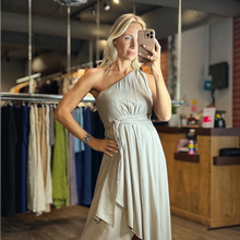 Load image into Gallery viewer, Model wearing a beige Aurora Boutique Miami one-shoulder flowy dress with a tie waist in a boutique setting.

