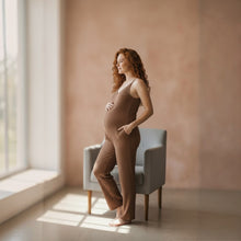 Load image into Gallery viewer, Pregnant model wearing a brown Aurora Boutique Miami cable knit jumpsuit with pockets, standing by a window.
