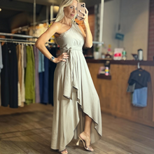 Load image into Gallery viewer, Model wearing an Aurora Boutique Miami cocoa one-shoulder flowy dress with a tie waist in a boutique setting
