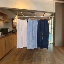 Load image into Gallery viewer, Aurora Boutique Miami Leonie linen pants in white, light blue, and navy hanging on a rack in a boutique.

